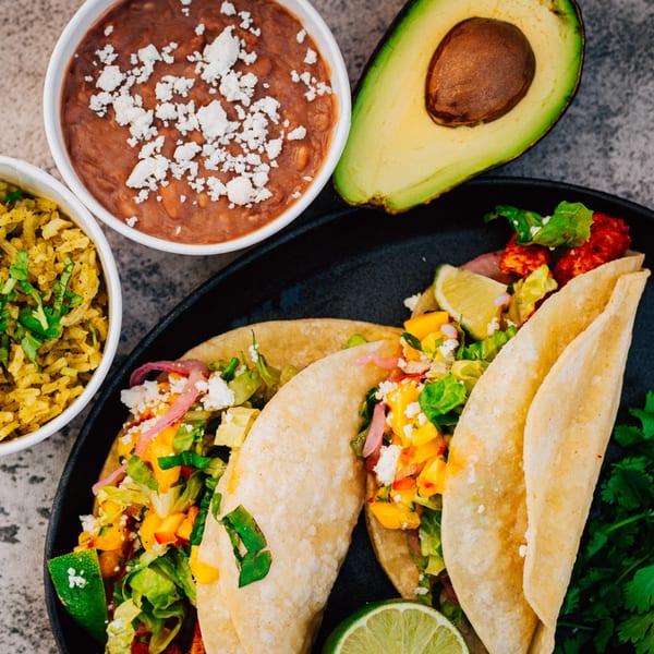 Three Serrano's Mexican tacos with rice, beans, and avocado on a dark plate.