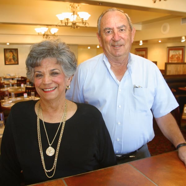 Smiling man and woman, likely owners, standing in a brightly lit Serrano's Mexican restaurant dining room.