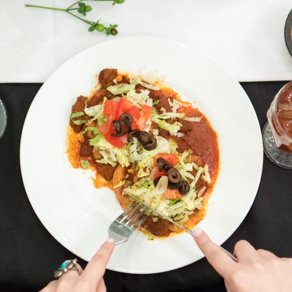 Overhead view of hands cutting into a plate of Serrano's Mexican enchiladas with lettuce, tomatoes, and olives.