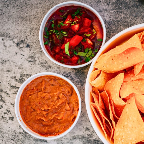 Overhead view of a bowl of red salsa, a bowl of refried beans, and a large bowl of tortilla chips.