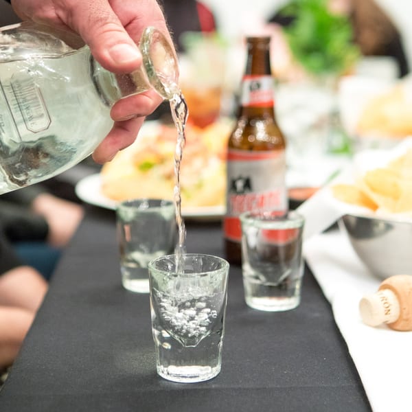 A hand pours clear liquid from a bottle into a shot glass, with other glasses and a beer bottle nearby.
