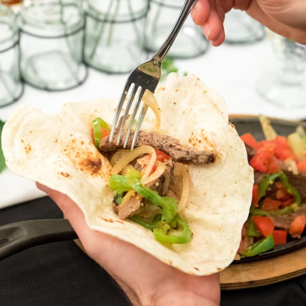 A hand holds a Serrano's fajita taco with steak, onions, and peppers, ready to eat.