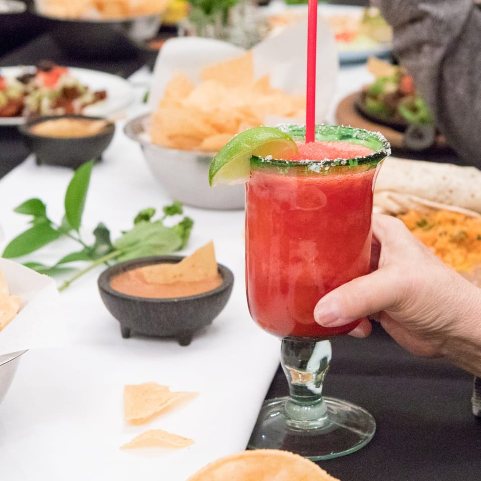 A hand holds a vibrant red strawberry margarita with a lime wedge and salted rim at Serrano's.